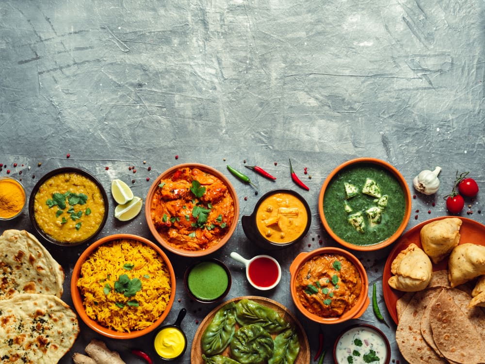 Small bowls of curries, dals and chutneys with spices on a blue stone table, seen from above